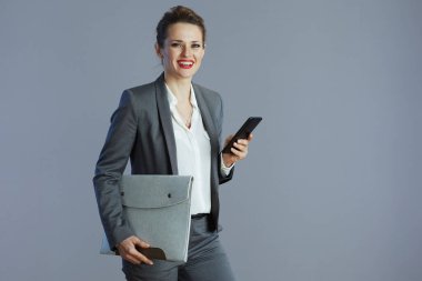 Gri arka planda izole edilmiş akıllı telefon ve klasörlü gri takım elbiseli, mutlu, küçük işletme sahibi kadın..