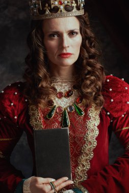 Kırmızı elbiseli ortaçağ kraliçesi, koyu gri arka planda kitap ve tacı var..