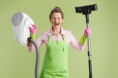 Bahar temizliği. Yeşil önlüklü mutlu orta yaşlı ev kadını ve yeşil elektrikli süpürgeye izole edilmiş lastik eldivenler..
