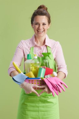 Bahar temizliği. Yeşil önlüklü ve lastik eldivenli mutlu modern kadın yeşil arka planda bir sepet temizlik ürünüyle izole edilmiş..