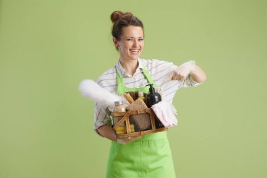 Bahar temizliği. Yeşil önlüklü mutlu orta yaşlı ev hanımı ve yeşil arka planda izole edilmiş lastik eldivenli çevre temizleme ürünleri sepeti..