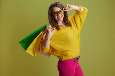 Sarı kazaklı, güneş gözlüklü, yeşil arka planda gülümseyen sağlıklı bir kadın..