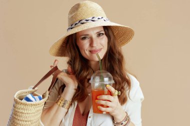 Plaj tatili Beyaz bluzlu, zarif orta yaşlı kadın ve bej arka planda saman torbası, havuç suyu ve yaz şapkası ile izole edilmiş şortlar..