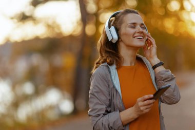 Merhaba sonbahar. Parkta spor kıyafetleri içinde gülümseyen şık bir kadın kulaklıkla müzik dinliyor..