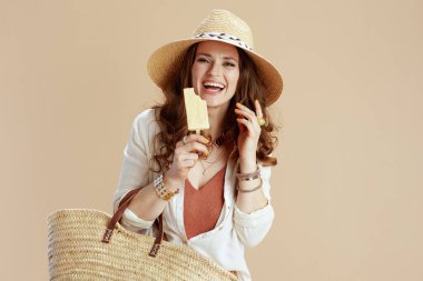 Plaj tatili Mutlu modaya uygun 40 yaşındaki beyaz bluzlu ve şortlu kadın bej arka planda saman torbası, dondurma ve yaz şapkalı..