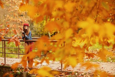 Merhaba sonbahar. Kot pantolonlu, kırmızı şapkalı, güz yapraklı, atkılı, eldivenli ve çantalı rahat modern kadın portresi..