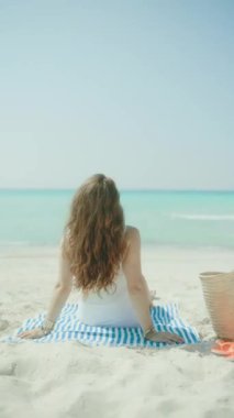 Rahat, modern, 40 yaşında bir kadın okyanus kıyısında saman torbası ve çizgili havluyla..