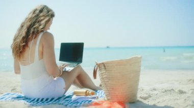 Okyanus kıyısında saman çantalı, dizüstü bilgisayarı boş ekranlı ve çizgili havlusu olan mutlu zarif bir kadın..