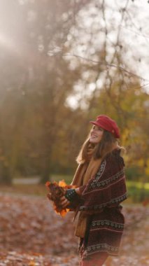 Merhaba sonbahar. Kırmızı şapkalı, eşarplı, eldivenli ve çantalı mutlu genç kadın şehir parkında neşe içinde..