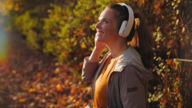 Merhaba sonbahar. Parkta spor kıyafetleri içinde, kulaklıkla müzik dinleyen mutlu modern bir kadın..