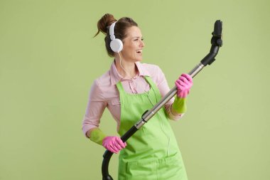 Bahar temizliği. Yeşil önlüklü, lastik eldivenli, yeşil arka planda izole edilmiş, elektrikli süpürgeli, gülümseyen 40 yaşında bir kadın..