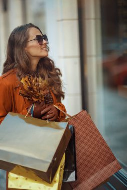 Merhaba Eylül. Şehirde alışveriş torbaları ve sonbahar sarısı yapraklarıyla turuncu trençkotlu mutlu orta yaşlı kadın..