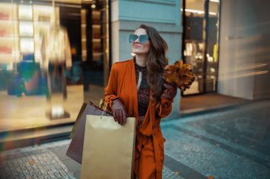 Merhaba sonbahar. Alışveriş torbaları ve sonbahar sarısı yaprakları olan turuncu trençkotlu modern kadın şehirde mağazanın yakınında..