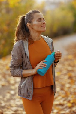 Merhaba sonbahar. Parkta spor kıyafetleri içinde, rahat, modern bir kadın ve bir şişe su..