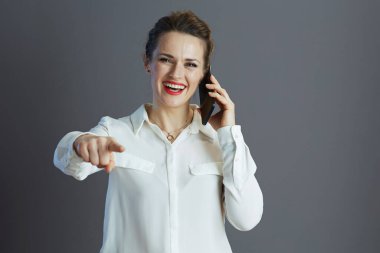 Beyaz bluzlu, güler yüzlü kadın çalışan kamerayı işaret ediyor ve gri renkli bir cep telefonuyla konuşuyor..