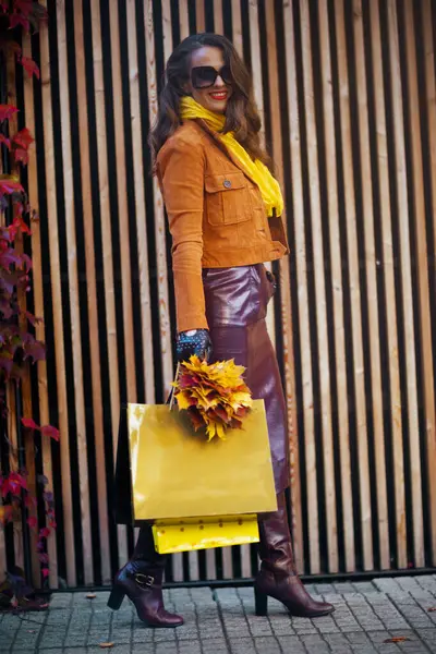 stock image Hello november. Full length portrait of smiling modern 40 years old woman in orange trench coat with shopping bags and autumn yellow leaves in the city.