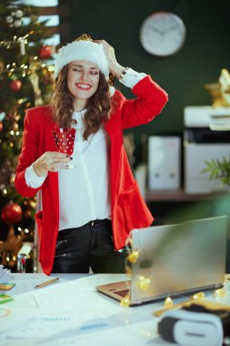 Noel zamanı. Kırmızı ceketle, Noel Baba şapkalı şarap kadehiyle modern yeşil ofiste Noel ağacıyla gülümsüyor..