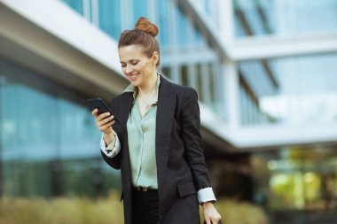 Orta yaşlı mutlu kadın işçi ofis binası yakınında siyah ceketli akıllı telefon kullanıyor ve yürüyor..