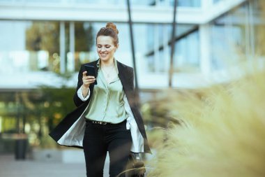 gülümseyen orta yaşlı kadın çalışan iş merkezinin yakınında siyah ceketli, evrak çantalı akıllı telefon kullanan ve yürüyen..