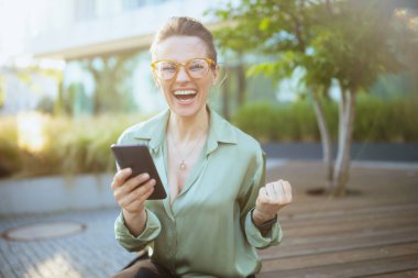 Yeşil bluzlu ve akıllı telefonlu gözlüklü, iş merkezinin yanında gülümseyen modern kadın işçi..