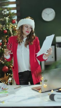 Noel zamanı. Gülümseyen orta yaşlı, Noel Baba şapkalı, kırmızı ceketli, modern yeşil ofisteki, Noel ağacı olan kırmızı ceketli, şık bir iş kadını..