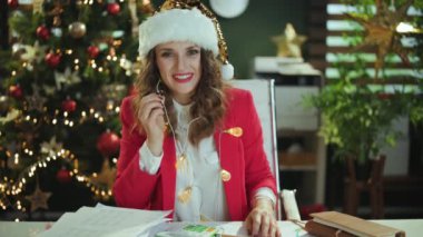 Noel zamanı. Noel Baba şapkalı, kırmızı ceketli, modern yeşil ofiste Noel ağacıyla video konferansı yapan orta yaşlı mutlu bir iş kadını..
