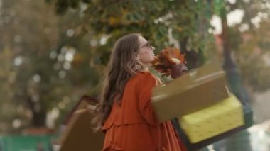 Merhaba Ekim. Alışveriş torbaları ve sonbahar sarısı yapraklarıyla kahverengi trençkotlu mutlu bir kadın..