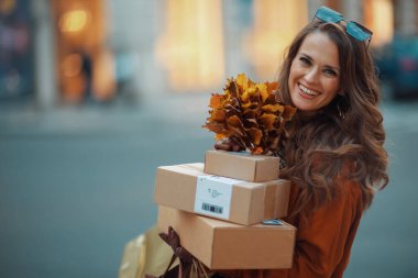 Merhaba Ekim. Kahverengi trençkotlu, paketli, güz sarısı yapraklı mutlu bir kadın..