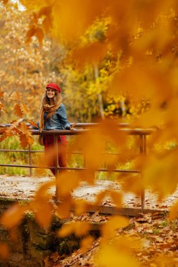 Merhaba sonbahar. Şehir parkında güz yaprağı ve eşarplı kot gömlek ve kırmızı şapkalı mutlu orta yaşlı kadın portresi.
