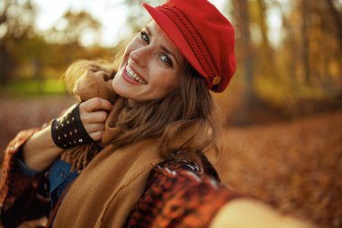 Merhaba sonbahar. Kırmızı şapkalı, eşarplı, gülümseyen zarif kadın şehir parkında video konuşması yapıyor..