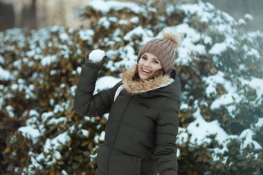 Şehir parkında kışın yeşil ceketli ve kahverengi şapkalı mutlu modern bir kadın. Eldivenleri ve karlı dallara kartopu atan bereli bir şapkası var..