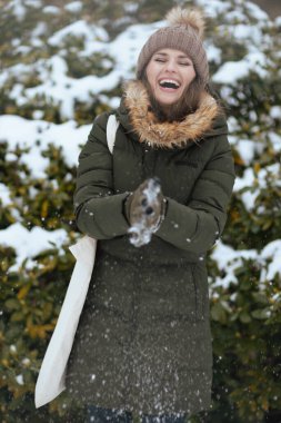 Güler yüzlü, yeşil paltolu ve kahverengi şapkalı modern kadın kışın şehir parkında eldivenleri ve karlı dalların yanında karla oynayan bere şapkalı..
