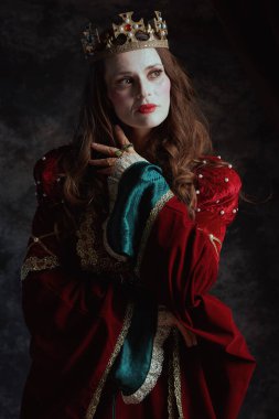 Kırmızı elbiseli, beyaz makyajlı ve tacı koyu gri arka planda olan ortaçağ kraliçesi..