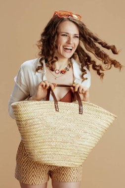 Plaj tatili Beyaz bluz ve şortla gülümseyen modaya uygun bir kadın saman çanta ve güneş gözlüğü ile bej arka planda.