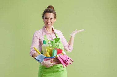 Bahar temizliği. Yeşil önlüklü, lastik eldivenli, yeşil arka planda izole edilmiş 40 yıllık mutlu modern kadın ve içi boş avuç içi temizleme ürünleri dolu bir sepet..