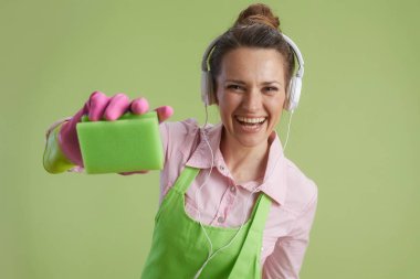 Bahar temizliği. Yeşil önlüklü ve lastik eldivenli gülümseyen modern kadın kulaklıkları ve temizleme süngeri ile yeşile izole edilmiş..