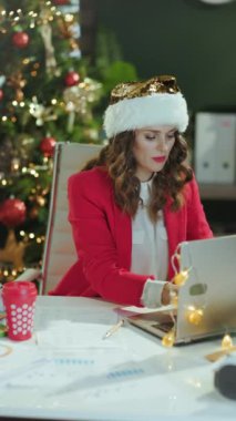 Noel zamanı. Noel Baba şapkalı, kırmızı ceketli, modern yeşil ofiste Noel ağacıyla çalışan mutlu ve zarif küçük işletme sahibinin portresi..