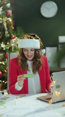 Noel zamanı. Gülümseyen, Noel Baba şapkalı ve kırmızı ceketli modern iş kadını. Laptop ve kredi kartıyla modern yeşil ofis ve Noel ağacı..