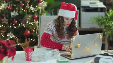 Noel zamanı. Noel Baba şapkası ve kırmızı Noel süveteri giyen, modern yeşil ofiste Noel ağacıyla çalışan yorgun, 40 yaşında bir iş kadını..