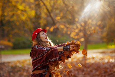 Merhaba sonbahar. Kırmızı şapkalı mutlu bir kadın. Eldivenli. Sonbahar yapraklarını şehir parkına atıyor..