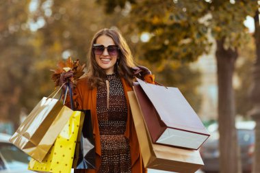 Merhaba Ekim. Turuncu trençkotlu, alışveriş çantalı ve sonbahar sarısı yapraklı gülümseyen genç kadın..