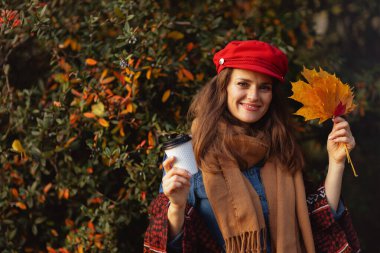 Merhaba sonbahar. Sonbahar yaprakları, atkısı ve kahvesi olan kırmızı şapkalı, gülümseyen genç bir kadın..