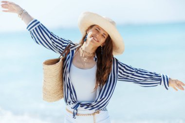 happy modern middle aged woman on the ocean shore with straw bag and straw hat rejoicing. clipart