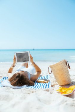 Okyanus kıyısında saman torbası ve çizgili havluyla tableti boş gösteren rahat ve zarif bir kadın..