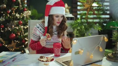 Noel zamanı. Noel Baba şapkalı, kırmızı kazaklı, geleneksel kurabiyeli, modern yeşil ofiste Noel ağacıyla sohbet eden zarif, küçük işletme sahibi kadın..