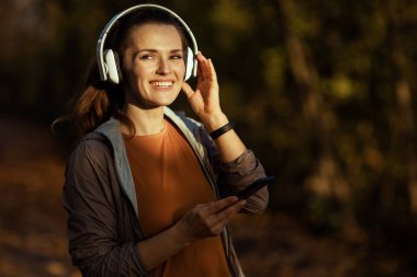 Merhaba sonbahar. Mutlu modern 40 yaşındaki kadın parkta kulaklıklı, spor elbiseli..