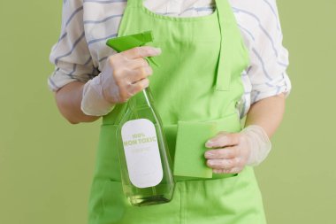 Bahar temizliği. Yeşil önlüklü, lastik eldivenli, yeşil arka planlı, çevre temizleme ürünü ve temizlik süngeri olan modern kadına yakın çekim..