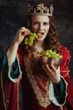 Kırmızı elbiseli ortaçağ kraliçesi. Üzüm tabağı ve tacı koyu gri arka planda..