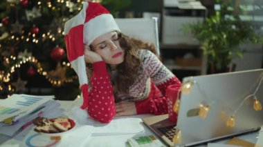 Noel zamanı. Noel Baba şapkalı, kırmızı Noel kazaklı, belgeli ve dizüstü bilgisayarlı, modern yeşil ofiste Noel ağacıyla uyuyan zarif bir iş kadını..