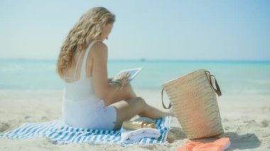 Rahat orta yaşlı kadın okyanus kıyısında saman torbası ve çizgili havluyla tablet bilgisayardaki uygulamaları kullanıyor..